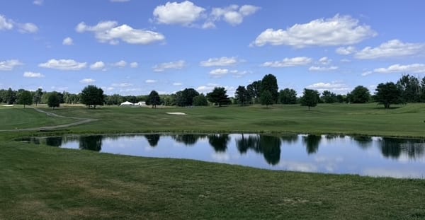 A pond reflecting the sky in front of open fairway and a distant tree line